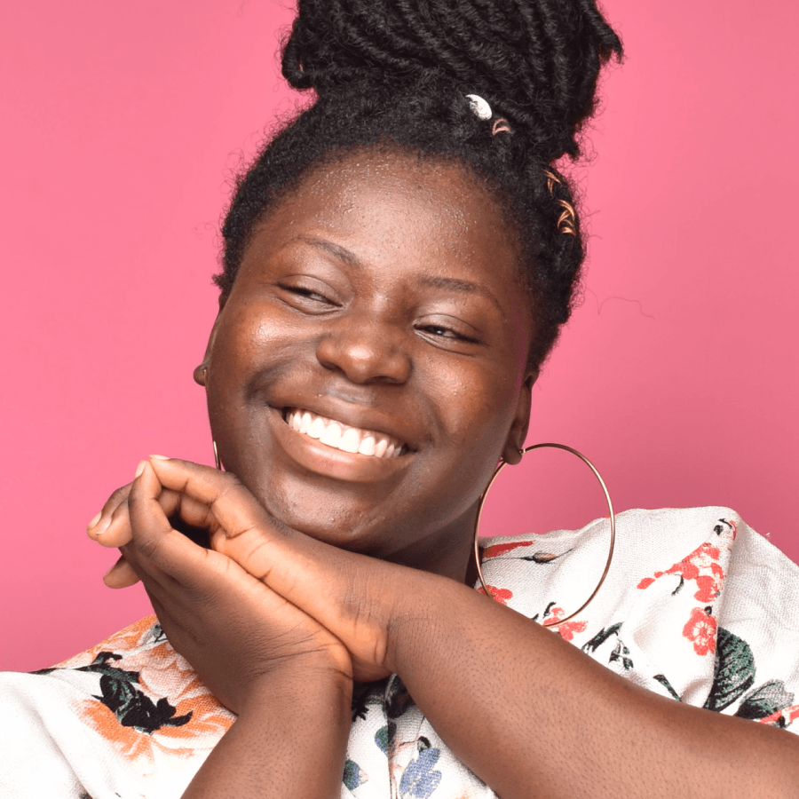 Jovem preta, de coque e blusa branca estampada, sorrindo com as mãos abaixo do queixo, olhando para o lado.