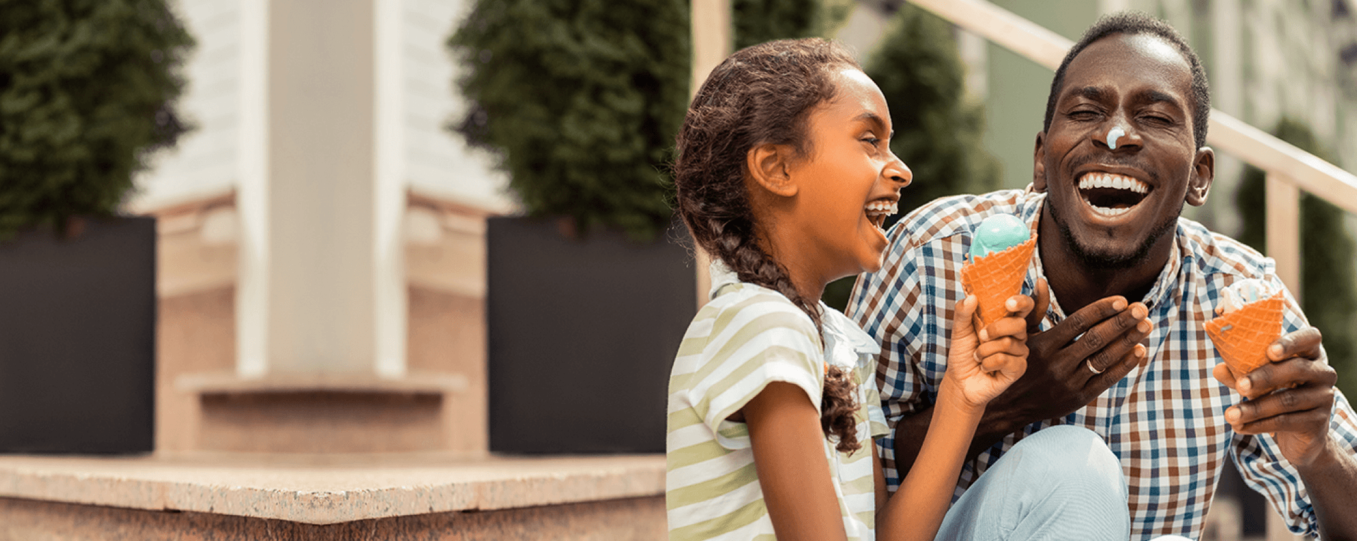 Homem preto e menina preta, sentados em uma escada, sorrindo enquanto seguram uma casquinha de sorvete na mão.