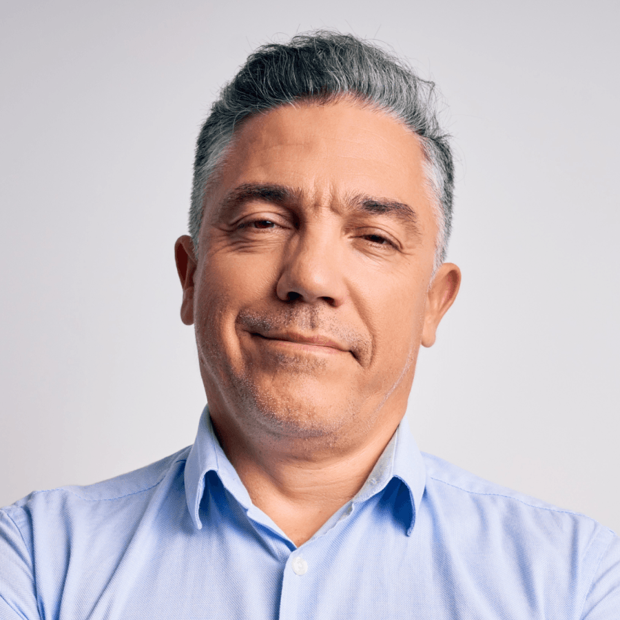 Homem branco, de cabelos grisalhos e topete, sorrindo de boca fechada olhando para a frente, de camisa azul claro.